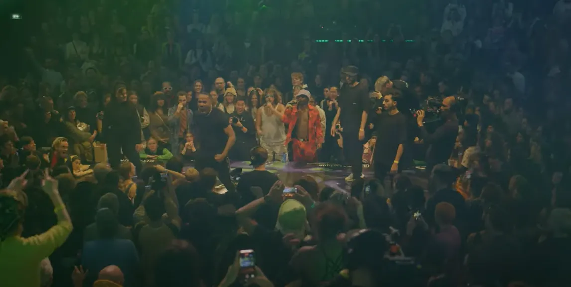 Crowd surrounding two pairs of dancers facing off at a dimly lit rave