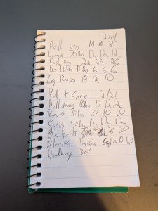 Small notebook with several exercises written on the lines with numbers of reps for sets written to the right